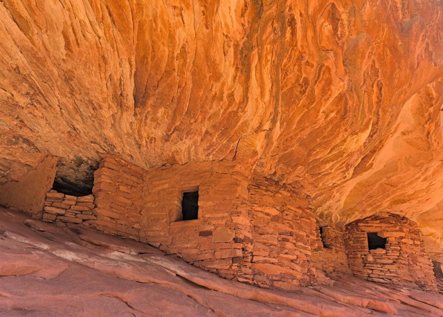 Color landscape photo of a cliff-dwelling. There's three rooms marked by three rectangular openings. The rooms are constructed of native orange-yellow sandstone blocks. The rooms a tucked under a orange-yellow sandstone cliff opening. The rock pattern above the rooms is streaks and looks like flames, like the rooms are on fire.