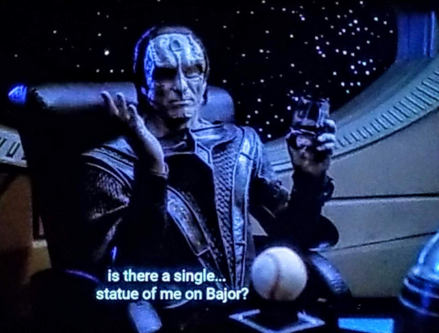 DS9 scene. Gul Dukat is sitting at a DS9 station desk, with a big space station window behind him showing stars. There is a baseball on the desk. Dukat is wearing a ridged hard leather official uniform that looks uncomfortable. His face and skin is gray, and he has boney ridges running all up and down his face, in one spot forming the shape of a spoon on his forehead. Kind of intimidating. He's holding his hands up like he's pondering something out loud. Closed caption reads, "Is there a single... statue of me on Bajor?"