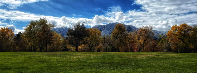 A panoramic view across a park, with an open expanse of green grass along the lower third of the photo, a line of trees in the middle with green, gold, orange, red, and brown autumn leaves, and the outline of mountains in the far distance behind the trees with a coating of fresh snow. A mix of broken clouds and blue sky above, with the sun trying to peek through the clouds on a chilly, autumn afternoon.