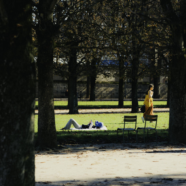 The image shows an urban park setting with a path surrounded by trees and foliage. In the foreground, a person is lying on the grass, seemingly resting or relaxing. A person in a yellow dress is standing nearby, facing the other direction. The scene has a peaceful, tranquil atmosphere.