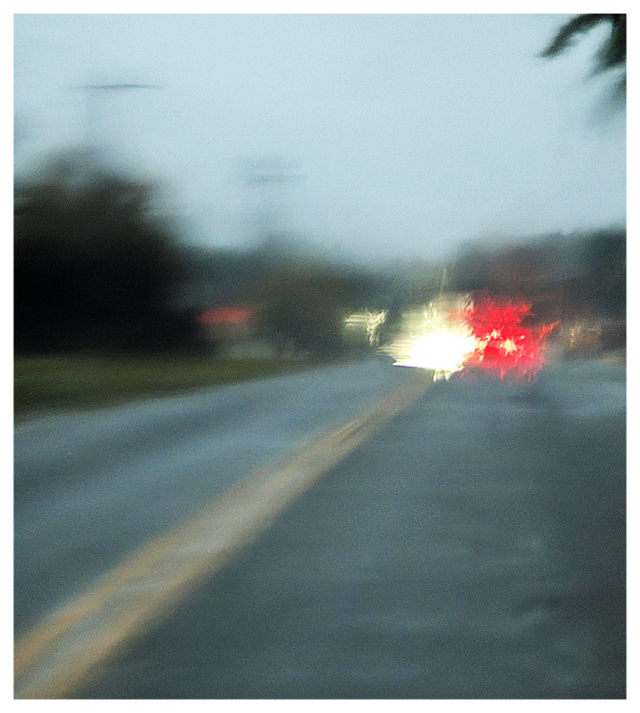 out of focus, two lane semi-rural road. rain. through a moving car windshield a car approaches, headlights on, a car brakes, taillights shine red. trees and grass. gray sky. 