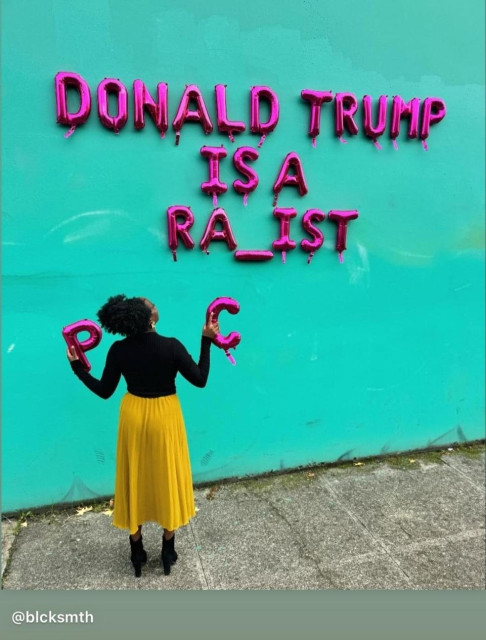 Color photo of a black woman in a gold skirt and black top holding two large pink letters, in her left hand, "P", in her right hand, "C". She's standing in front of a blue wall looking up at this phrase in large pink letters, "DONALD TRUMP IS A RA_IST".