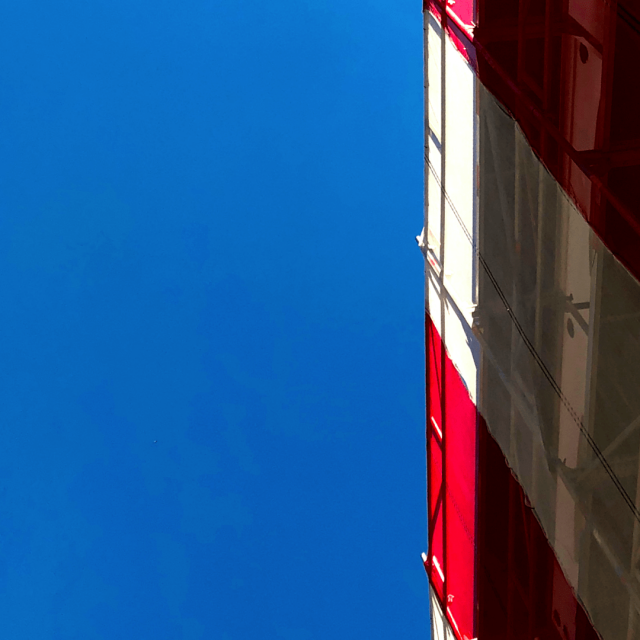 On the far right of the frame is a tarp with large red and white stripes covering a scaffold. In the center and left of the frame is a clear blue sky.