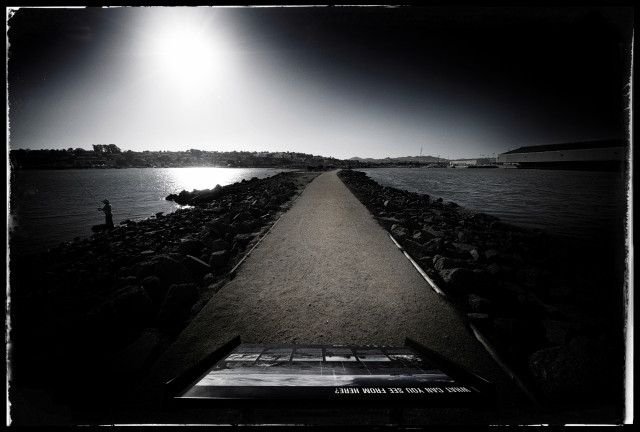 A dark scene of a dirt path down the center of a spit of land that juts out into San Francisco Bay. On either side of the path: rocks and water. On the left, a fisherman. On the far right, a long shed that houses a recycling plant. Above: a glaring, angry sun that has caused my camera's metering to go crazy.

In front: a park sign... a guide to the area that asks "what can you see from here?"