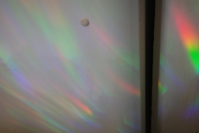 A close-up view of a wall with a subtle texture, featuring colorful rainbow light reflections and a small round object on the surface.