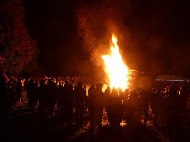 A large, blazing bonfire at night, with a circle of people standing around it.