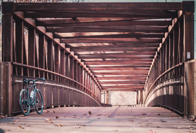 Photo of a black specialized diverge gravel bike standing at the entrance of a metal footbridge. The bridge is rust colored, and has a slight arch to it. It has a roof made of crisscross metal beams. We are looking past the bike into the bridge and over the arch and at the end of the bridge, where there’s a small opening showing a tree with autumn leaves on it. 