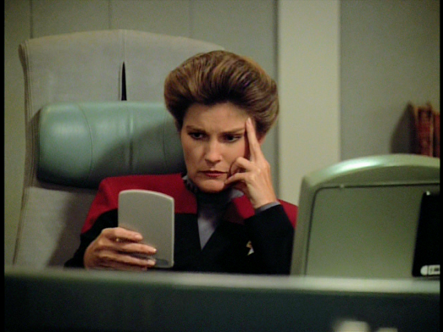 Janeway, like all of us, sits at her desk, staring intently at a small device in her hands. 