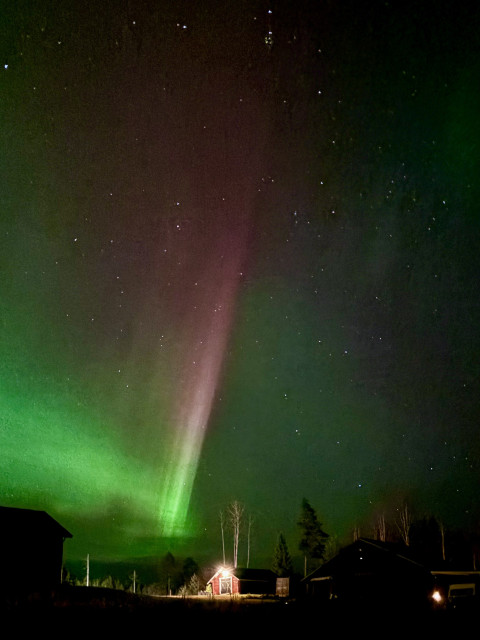 A bright red and green Aurora shooting across the sky above some outbuildings.