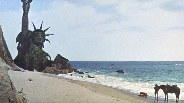 A weathered statue of Liberty is partially submerged on a beach, with waves lapping at the shore. In the foreground, two figures are seen, one interacting with a horse, while the ocean stretches into the background under a cloudy sky. The final scene from the iconic Planet of the Apes starring Charlton Heston.