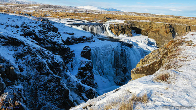 A photo of a landscape. In the centre of the shot is a pretty waterfall with multiple small drops, most of them seemingly completely frozen. In reality the water is still flowing beneath the ice but that isn't apparent from this shot. The surrounding rock surface is honey-coloured, the vegetation is brown and withered and the horizontal surfaces are covered in snow. The low light from the left is casting long shadows across the falls making the water and ice seem blue, and the brown rock almost black.