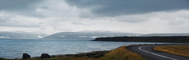 A wide photo of a landscape under grey cloud. The foreground is brown vegetation and a tarmac road is looping from lower right to centre right. Beyond it is a body of ruffled turquoise water. In the misty distance are snow-striped headlands, on the left are two mountains with a deep valley between them. The whole scene is peaceful, and calming.