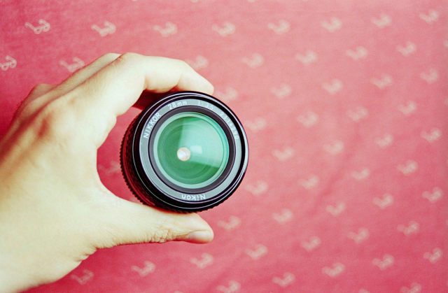 Lens in hand with the glass pointing at the viewer on a pink textured background