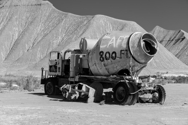 Black and white landscape photo of a derelict cement truck sitting in a barren field and barren heavily erode small angular hills behind it. The truck is pointing toward the hills on the left. The driver's door is open and the tires are flat. A sign is painted on the large cement mixer cylinder that says, Cafe 800ft."