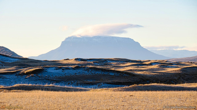 A photo of a landscape under a clear sky. The foreground is withered brown grassland, patches of hard frost are visible in the deep shadows. In the distance is an unusual mountain. It has a pointed summit which tapers slightly to form a table top. From there the shoulders of the peak are steep, gradually widening in a soft curve to ground level. There's a tuft of bearded cloud sitting on the top. Other mountains can be seen in the distance, fading with tonal perspective in the deep cold. It's very beautiful.