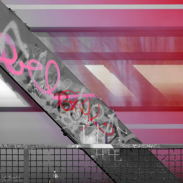 A steel beam runs diagonally from top left to bottom right through the picture. Graffiti and writing can be seen on the steel beam. Behind it, a train passes through, which is blurred by a longer exposure time. Only the colors pink, red and purple remain in the picture. "THE BASTARDS" is written on a railing.