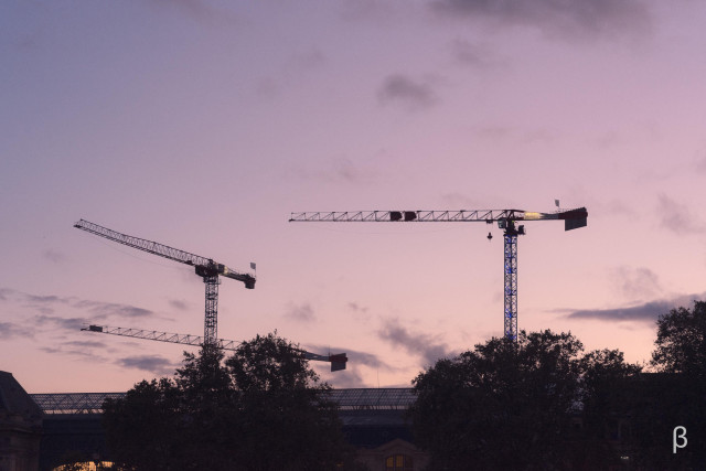 The image shows several construction cranes silhouetted against a dramatic, colorful sky at sunset. The cranes are tall, dominant structures in the frame, suggesting an ongoing construction project or development. The clouds in the sky have a vibrant, pinkish-purple hue, creating a striking contrast with the dark silhouettes of the cranes. The overall scene has a moody, atmospheric quality, highlighting the interplay between the industrial, man-made elements and the natural beauty of the sky.