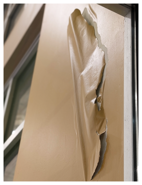 low angle view through a glass supermarket window of a beige wall with a large, vertical tear in the paint. out of focus windows at left. 