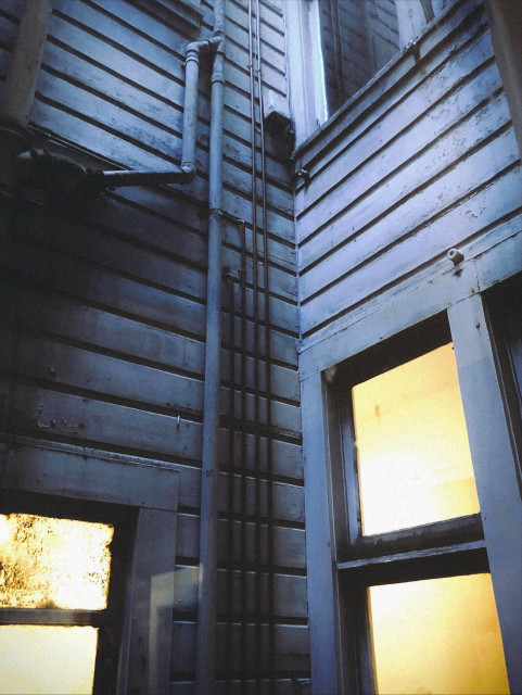 A grim photo of a moody San Francisco lightwell scene. We are looking out one window into a small contained space between buildings, onto a few musty yellow-stained windows lit from within, and a network of utility pipes and wires climbing up the bluish-grey, wooden-clad horizontal house siding. Very ominous and foreboding. 