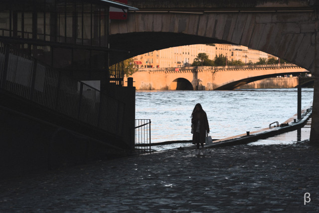 The image shows a person standing alone beneath a bridge along the Seine River in Paris. They are silhouetted against the warm, golden light that illuminates the historic buildings and another bridge visible through the arch. The person appears to be carrying a shopping bag and is positioned on a wet riverbank walkway. The contrast between the dark underpass and the bright, sunlit scene beyond creates a dramatic framing effect, capturing a contemplative moment in the urban landscape.