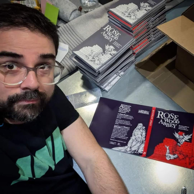 Selfie of Kalum, a bearded white man with glasses, in front of a table covered with copies of Rosewood Abbey and dust covers awaiting to be folded.