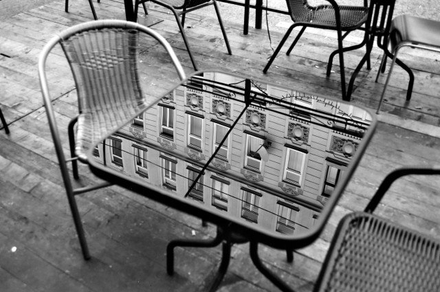 A black-and-white photograph of an outdoor cafe scene, where a reflective tabletop captures the detailed reflection of an ornate building facade with windows and decorative elements. The chairs surrounding the table are empty, and the polished table surface sharply mirrors the architecture above, creating a striking juxtaposition between the reflection and the wooden deck below.