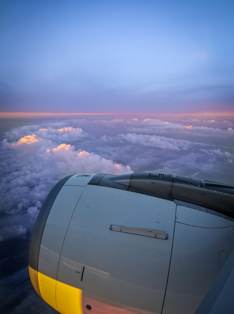 A sea of orange and purple clouds shot from the window of a plane, the front of the engine glowing yellow as it catches some sunlight.