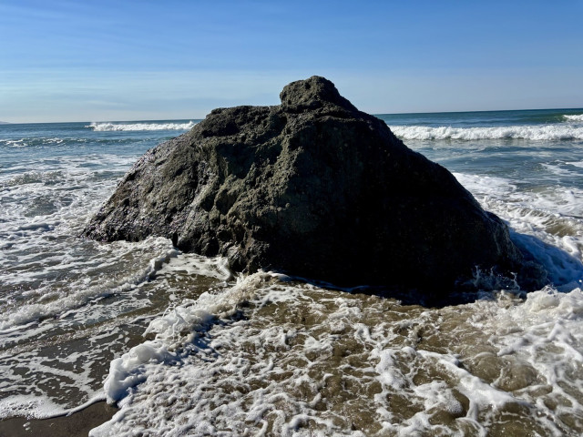 Cayucos, CA. A large rock partially submerged in shallow water, with waves crashing around it. The background features a clear blue sky and distant ocean waves.