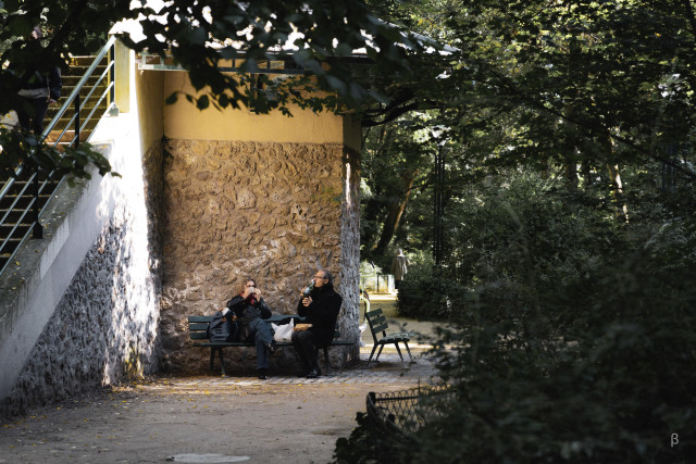 The image shows two people sitting on a bench in a sheltered stone alcove or nook, surrounded by greenery and trees. They appear to be taking a break or having a meal, with one person holding what looks like a drink or food item. The scene is peaceful and intimate, with dappled sunlight filtering through the leaves and creating shadows on the stone walls. The architecture appears to be part of a park or garden setting, with a rustic, European character.