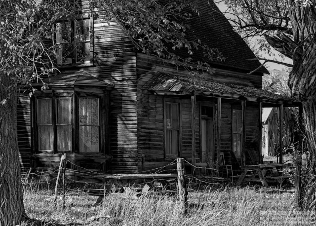 Black and white landscape photo of a very old weathered two story wood house. The image is framed with parts of tree trunks visible on each side. Between the trees the old house is visible. The view is toward the right corner of the house with a covered porch and front door flanked by a window on either side on the right side of the frame. The right side of the house (left side of image) shows a nook and upper floor window. The roof is very steep. In the frontyard, just beyond the porch, is a wood picnic table with benches. There's a falling down barbed wire fence with wooden fence posts running across the image between the trees and the house.