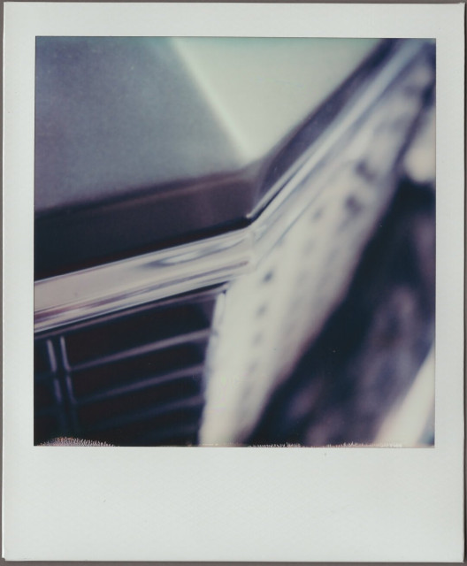 Polaroid of the front grill and hood of a gray, '70s Buick Riviera, all very monochrome and angular.