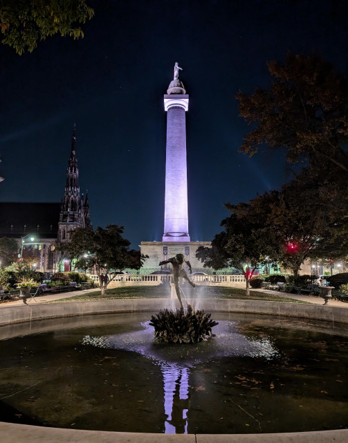 Washington Monument (Baltimore) lit up in purple and reflected in a fountain in the foreground