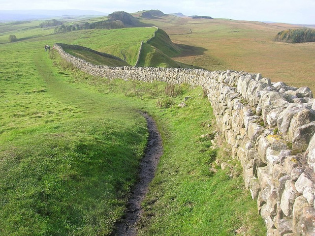 Photograph of a section of Hadrian’s wall which shows it following a ridge line. There’s a path next to it where visitors can walk along that has been created by many feet walking a similar line.