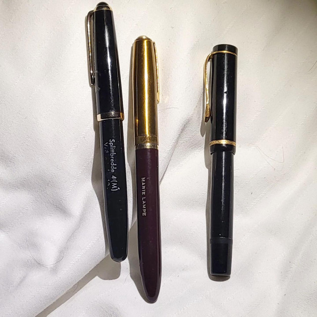 Three capped fountain pens on a white background. The center is maroon with a goldtpnencap, the other two are black with gold trim.