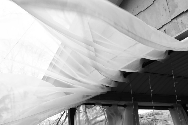 A monochrome image featuring flowing sheer curtains inside a structure, with visible light filtering through. The curtains create a soft, ethereal atmosphere against a backdrop of a partially obscured exterior.