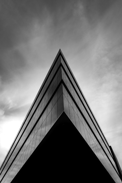 Fotografía en blanco y negro donde se ve un edificio de arquitectura actual acristalado tomado en contrapicado. El cual muestra una forma triangular afilada como en forma de flecha sobre un cielo difuminado.