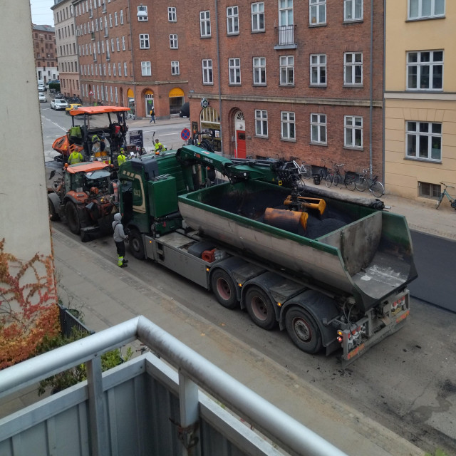 Construction crew paving the street - there's a big truck half-emptied of its load of asphalt, some other machinery and guys in workwear. The front of the scene is the corner of my balcony and the backdrop is red and yellow Copenhagen apartment buildings