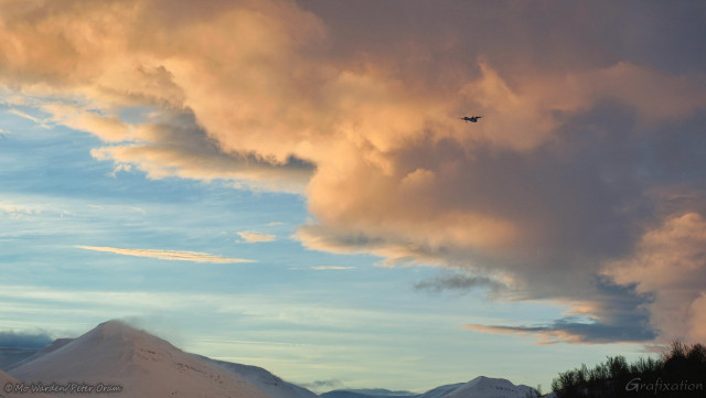 A photo of a cloudy sky with snow-capped mountains in the lower part of the shot. In the top right quarter is a small aircraft, two propellers are visible. The clouds behind it are lit by the rising sun with tones of orange and yellow, and there is heavier and darker cloud behind it but most of the sky is quite clear. The mountain tops are displaying spindrift snow from the summits, a sign of strong winds.