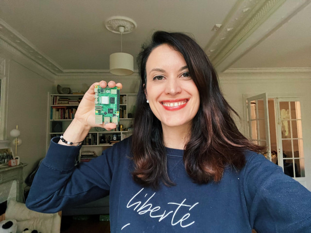 A selfie showing your truly (Caucasian woman with long brown hair) holding up a Raspberry Pi and smiling to the camera. I am wearing a blue sweatshirt that says “liberté” (freedom) 