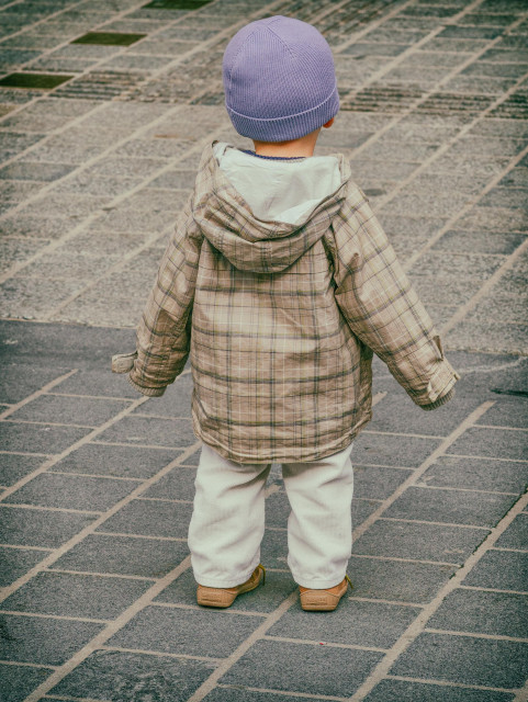 Ein kleiner Bub mit heller Hose, karierter Jacke und einer fliederfarbenen Mütze steht da ganz allein. Man sieht nur den Rücken des Buben.
Das Bild ist in Farbe und im Querformat aufgenommen.