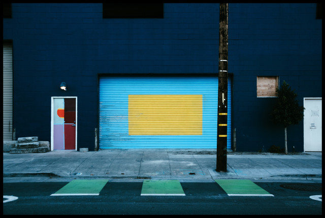 A photo of the side of a building that caught my eye because of the colorful, whimsical paint job. Not quite a mural and a few boxes short of a Mondrian... the building's dark blue wall, the door, and the large garage door are painted with rectangles of seemingly random colors (light blue, yellow, lavendar, maroon...). Similarly, a lane on the street in front has green rectangles that jut out from the sidewalk.

Art.