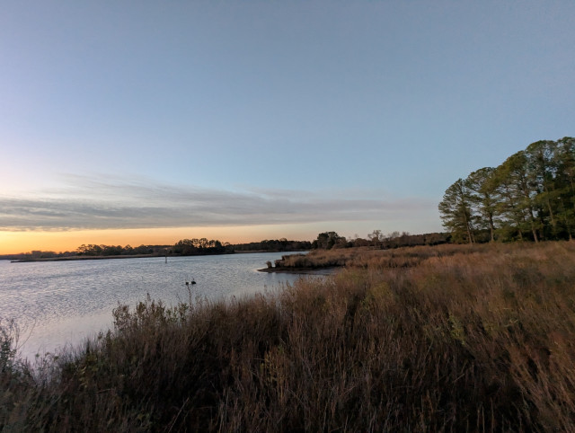 Photo of a calm morning sunrise over Harris Creek sun rising below the horizon and tinting it orange. Streaks of grey stratus hang in the sky to the right.
 Marsh is brown and ready for a long winter nap. Creek water is calm and perfectly reflects the sky and clouds.