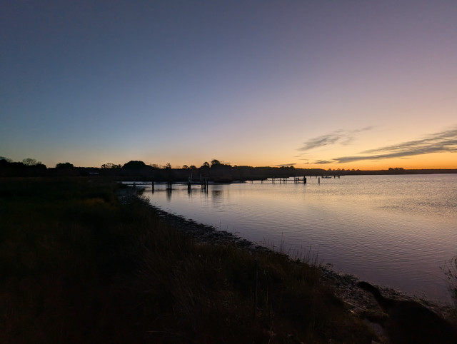 Photo of a calm morning sunrise over Harris Creek sun rising below the horizon and tinting it orange. Streaks of grey stratus hang in the sky to the right.
 Marsh is brown and ready for a long winter nap. Creek water is calm and perfectly reflects the sky and clouds.