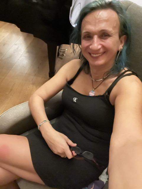 Selfie of me, a non-binary person, wearing a black spaghetti strap mini dress, sitting on a chair and smiling into the camera. I look very happy and relaxed.