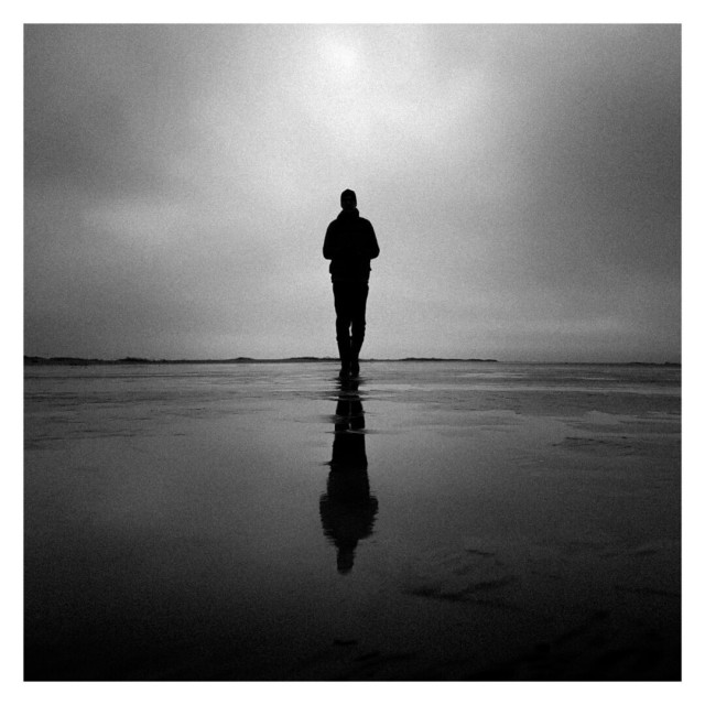 The image is a square black-and-white photograph with a white border, depicting a solitary figure standing on a vast, reflective surface that seems to be a wet, flat beach or a shallow body of water. The person is silhouetted against a soft, overcast sky, creating a striking contrast. Their reflection is visible in the water beneath, elongating the figure in a mirror-like effect that adds depth to the scene. The horizon line is faint, blurring the boundary between sky and land, lending a sense of infinite space. The overall mood of the photo is contemplative and somber, with the minimal composition highlighting the lone figure in a vast, empty environment. The subdued light and grayscale tones contribute to a feeling of solitude and introspection.