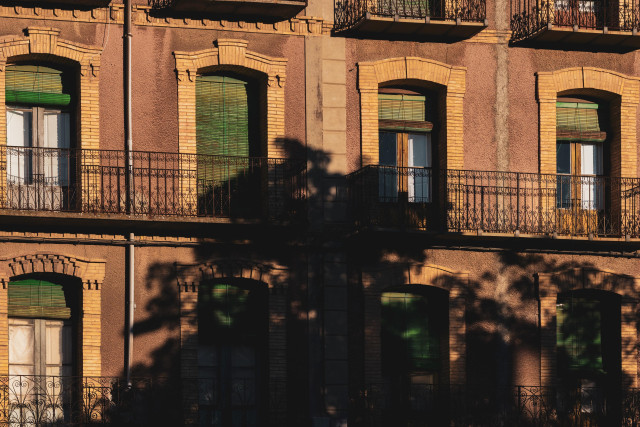 Se ve una fachada con dos hileras de con balcones y cuatro ventanales por hilera. En la parte central, ocupando bastante espacio, la sombra oscura y marcada de un árbol por la luz crepuscular de la tarde.