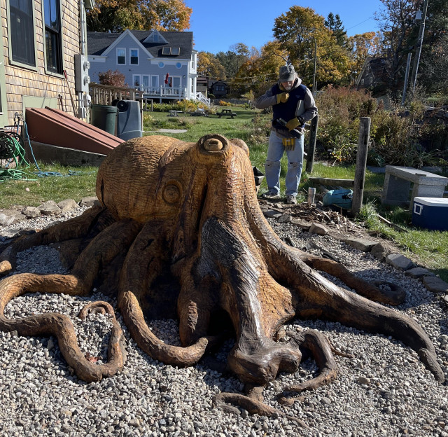 In the foreground is an octopus sculpted out of a tree stump. In the background is the local sculptor who created this work of art. He is wearing a baseball cap and work gloves and looking down. 