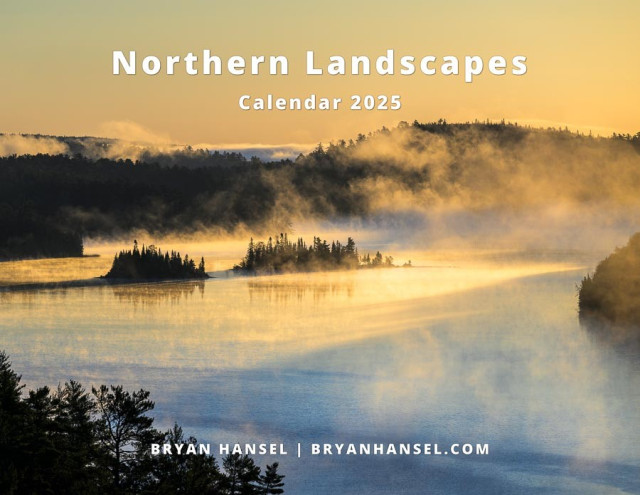 A calendar cover showing a lake scene surrounded by hills. The lake has two islands on it and mist and fog is floating through the air. It's early morning so the sun is turning everything yellow and casting shadows across the lake. The shadows are blue. The cover is titled "Northern Landscapes Calendar 2025." At the bottom, it shows the author "Bryan Hansel | bryanhansel.com."