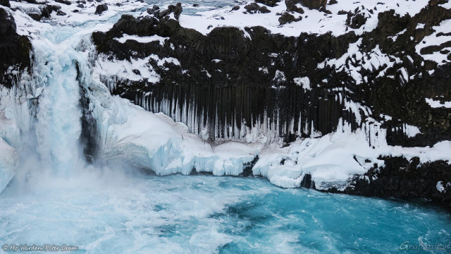 A photo of a horsetail waterfall on the left of a frozen landscape. The water in the pool below the chute is turquoise and foam is churning across the surface. The cascade is surrounded by near vertical columns of black basalt, and the white ice between them is highlighting the joints in the structure. Around the base, the spray has created large blocks of cyan ice capped with snow. The crest of the cascade is over-topped by a delicate arch of ice, and the river feeding the falls can be seen at the top left to centre of the shot.