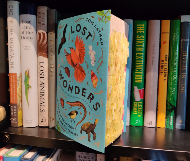 A photo of Tom Lathan's new book Lost Wonders, tilted towards the viewer such that the large number of yellow post-it notes show. The book is standing on a black bookshelf full of books about (mass) extinctions.